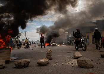 Burning vehicles and thick black smoke on a city street during a protest or riot, with people observing and taking photos.