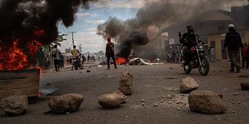 Burning vehicles and thick black smoke on a city street during a protest or riot, with people observing and taking photos.