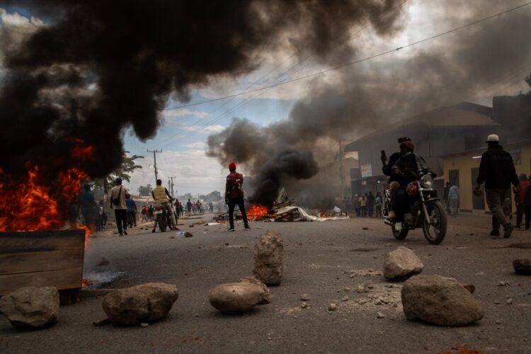 Burning vehicles and thick black smoke on a city street during a protest or riot, with people observing and taking photos.