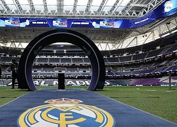Aerial view of a modern football stadium prepared for a match, featuring the Real Madrid logo on the pitch and a circular tunnel for players to enter the field. The stadium is equipped with large screens and bright lighting for an exciting sporting event.