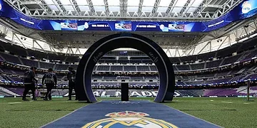 Aerial view of a modern football stadium prepared for a match, featuring the Real Madrid logo on the pitch and a circular tunnel for players to enter the field. The stadium is equipped with large screens and bright lighting for an exciting sporting event.