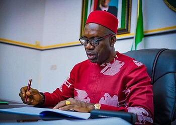 African man in traditional attire signing documents at a desk in an official setting with flags in the background.