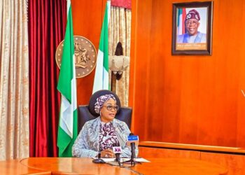 African woman government official speaking at a press conference with Nigeria flags in the background.