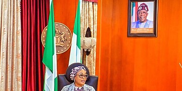 African woman government official speaking at a press conference with Nigeria flags in the background.