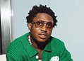 A young man with curly hair wearing sunglasses and a green sweater, looking confidently at the camera.