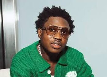 A young man with curly hair wearing sunglasses and a green sweater, looking confidently at the camera.