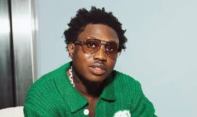 A young man with curly hair wearing sunglasses and a green sweater, looking confidently at the camera.