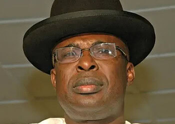 Photograph of a man wearing a black hat and glasses, looking serious in an indoor setting.