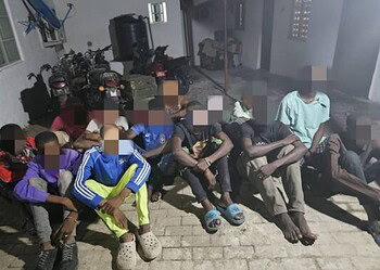 Innocent children sitting outdoors on a sidewalk, some with their hands behind their backs, during daytime, with motorcycles and a building in the background.
