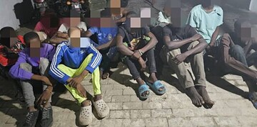 Innocent children sitting outdoors on a sidewalk, some with their hands behind their backs, during daytime, with motorcycles and a building in the background.