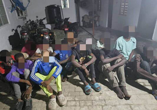 Innocent children sitting outdoors on a sidewalk, some with their hands behind their backs, during daytime, with motorcycles and a building in the background.