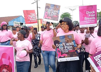 Breast cancer awareness march with women holding signs promoting early detection and health checkups, wearing pink outfits on a street.