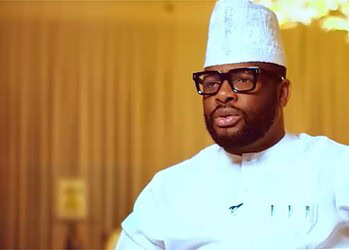 Professional Nigerian chef wearing traditional attire, glasses, and a hat, speaking confidently in a modern kitchen setting.