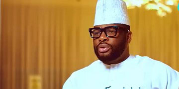Professional Nigerian chef wearing traditional attire, glasses, and a hat, speaking confidently in a modern kitchen setting.