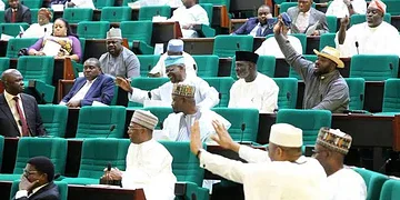 Seated lawmakers and officials engaged in a parliamentary debate in a legislative chamber with green seats.