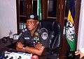 Police officer in uniform Ondo Prepaid Meter Arson case undersmiling at desk in an office with flag of Sierra Leone in background.