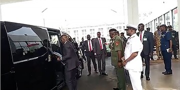Black luxury vehicle being attended to by a man in a suit, surrounded by distinguished military and civilian officials at an official event, possibly a formal or diplomatic occasion.