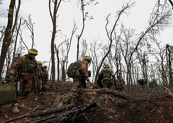 Soldiers in camouflage gear navigating a forested area affected by fire, carrying supplies and equipment, demonstrating military deployment in disaster or conflict zones.