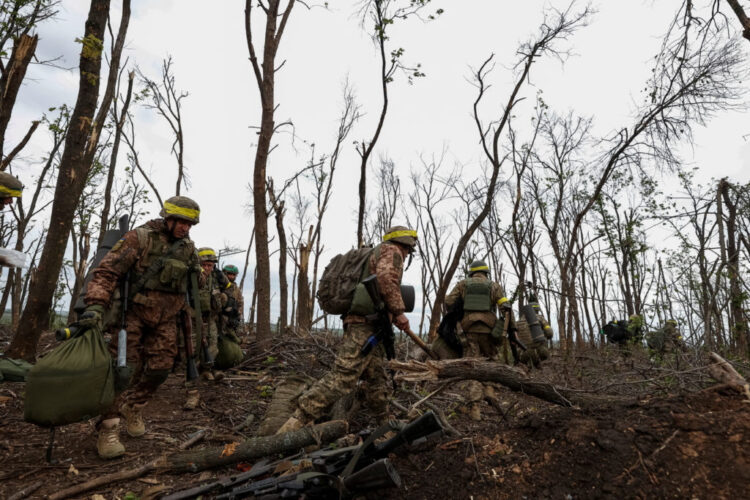 Soldiers in camouflage gear navigating a forested area affected by fire, carrying supplies and equipment, demonstrating military deployment in disaster or conflict zones.