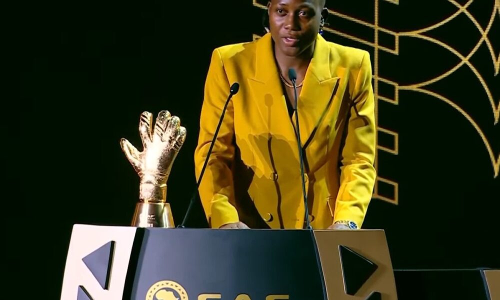 Bold woman in a yellow blazer presenting at the Freelanews event, standing behind a podium with a trophy and speaking into microphones, highlighting recognition and achievements in freelancing or recognition awards.
