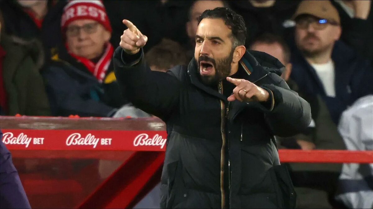 Assertive football coach passionately shouting and pointing during match, surrounded by cheering fans, behind Bally Bet advertising at football stadium.
