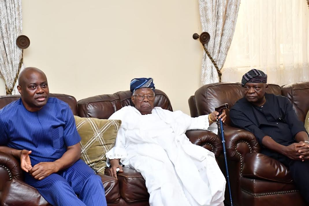 Elderly man in traditional white attire sitting on leather sofa with two men in modern attire, all in a formal living room setting, representing Nigerian cultural and political heritage.