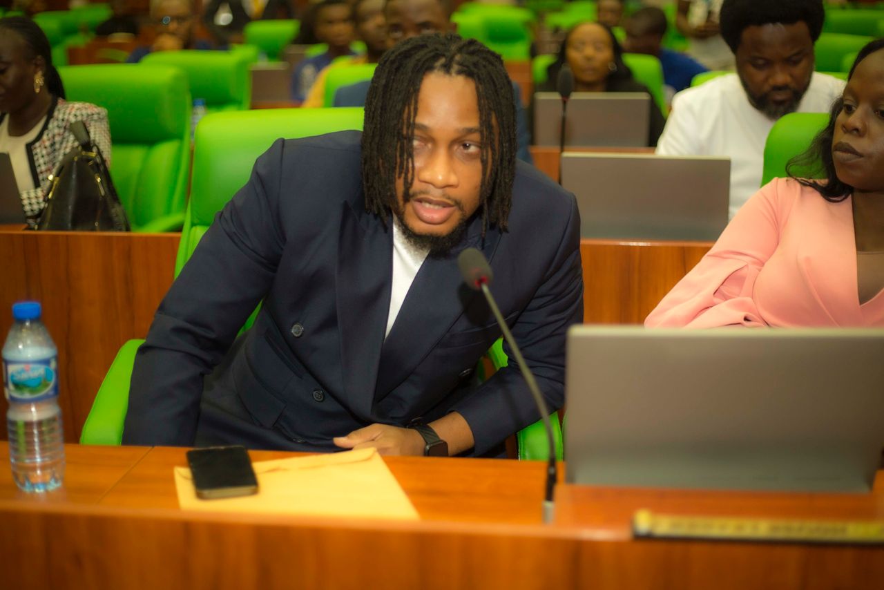 A young man in a navy suit speaking at a conference or seminar, surrounded by attendees in a professional setting with green chairs.