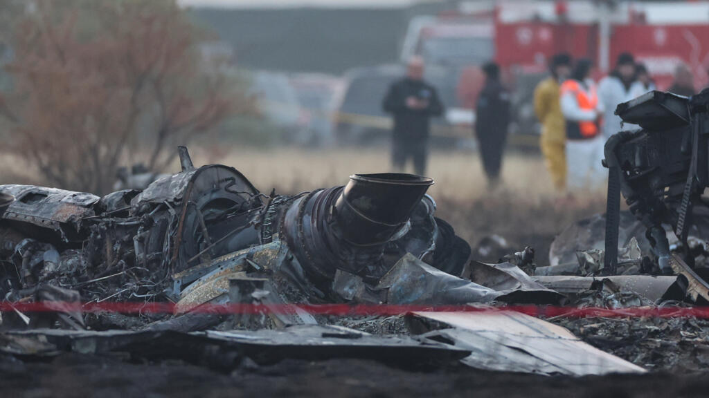Damaged aircraft wreckage from a fatal crash scene, with emergency responders in the background attending the site.