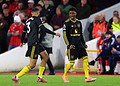 Dynamic football players celebrating a goal during a match, wearing black and yellow team kits, on a well-lit stadium with enthusiastic spectators in the background.