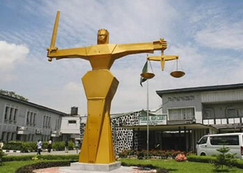 Majestic golden statue of Lady Justice holding scales and a sword, symbolising fairness and law, situated outdoors near a modern government building with a clear blue sky.