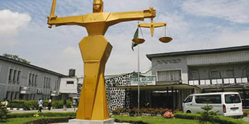 Majestic golden statue of Lady Justice holding scales and a sword, symbolising fairness and law, situated outdoors near a modern government building with a clear blue sky.