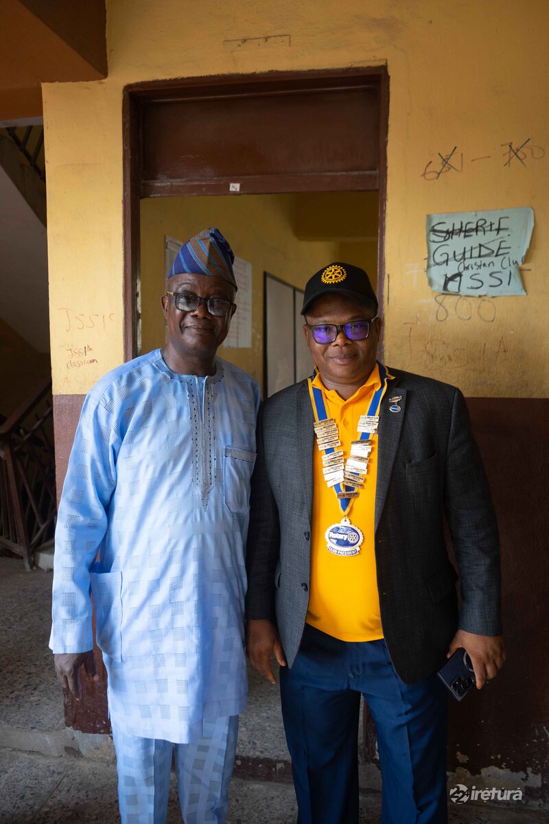 A man wearing a grey blazer with a Rotary International badge and a yellow polo shirt stands next to a man in traditional blue attire with a cap, both posing inside a building, showcasing community leadership and local engagement.