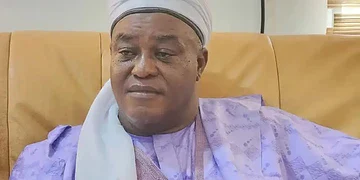 Elderly man wearing traditional attire and headwrap sitting on a beige sofa in a well-lit room, showcasing diverse cultural fashion.
