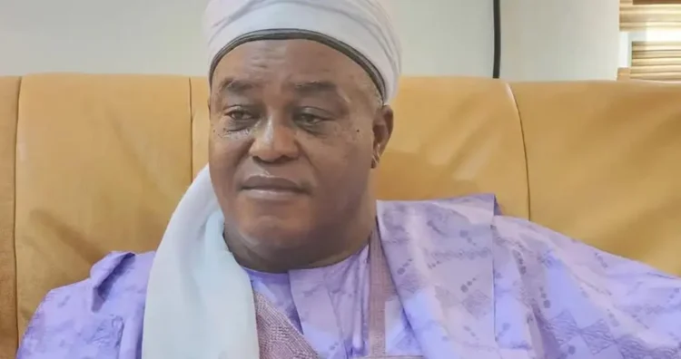 Elderly man wearing traditional attire and headwrap sitting on a beige sofa in a well-lit room, showcasing diverse cultural fashion.