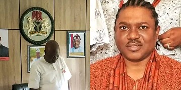 A government official in a white outfit standing in an office with framed photos and emblems on the wall, representing official political or administrative context in Nigeria.
