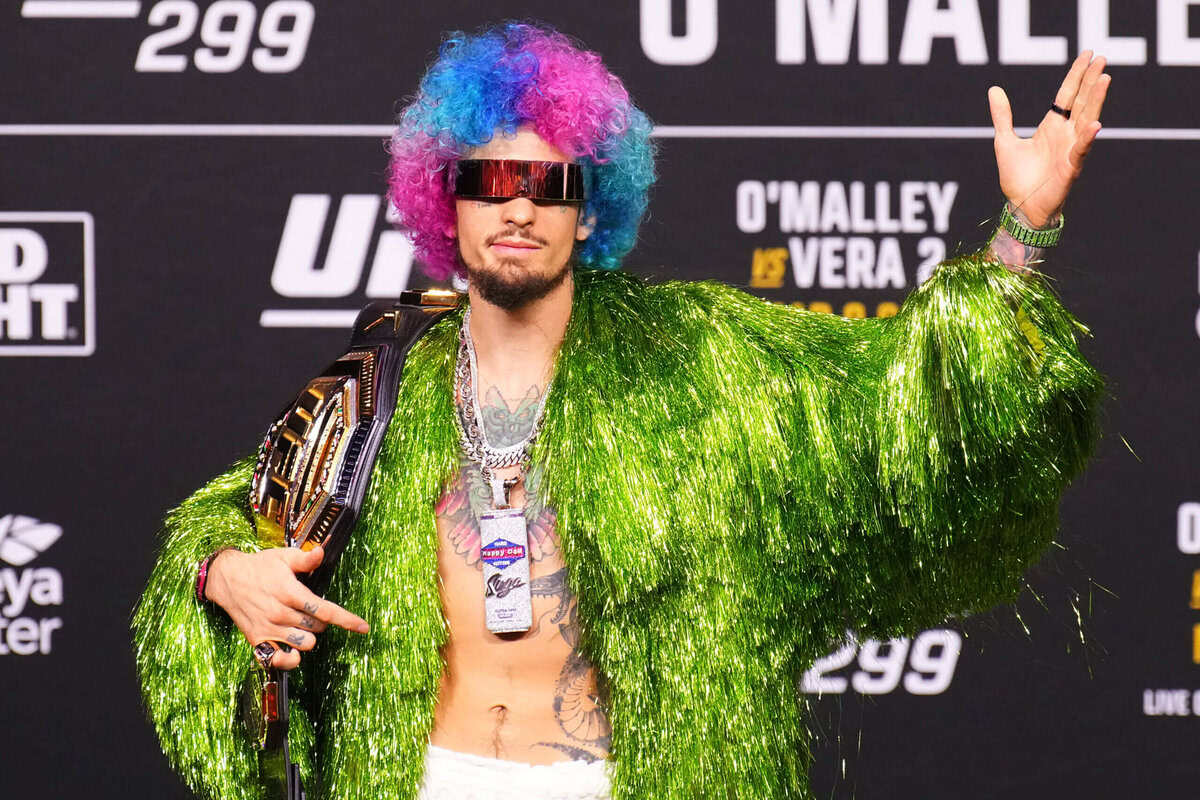 A colourful UFC fighter in a shiny green jacket and futuristic visor, showing his championship belt at a weigh-in event with promotional banners in the background.