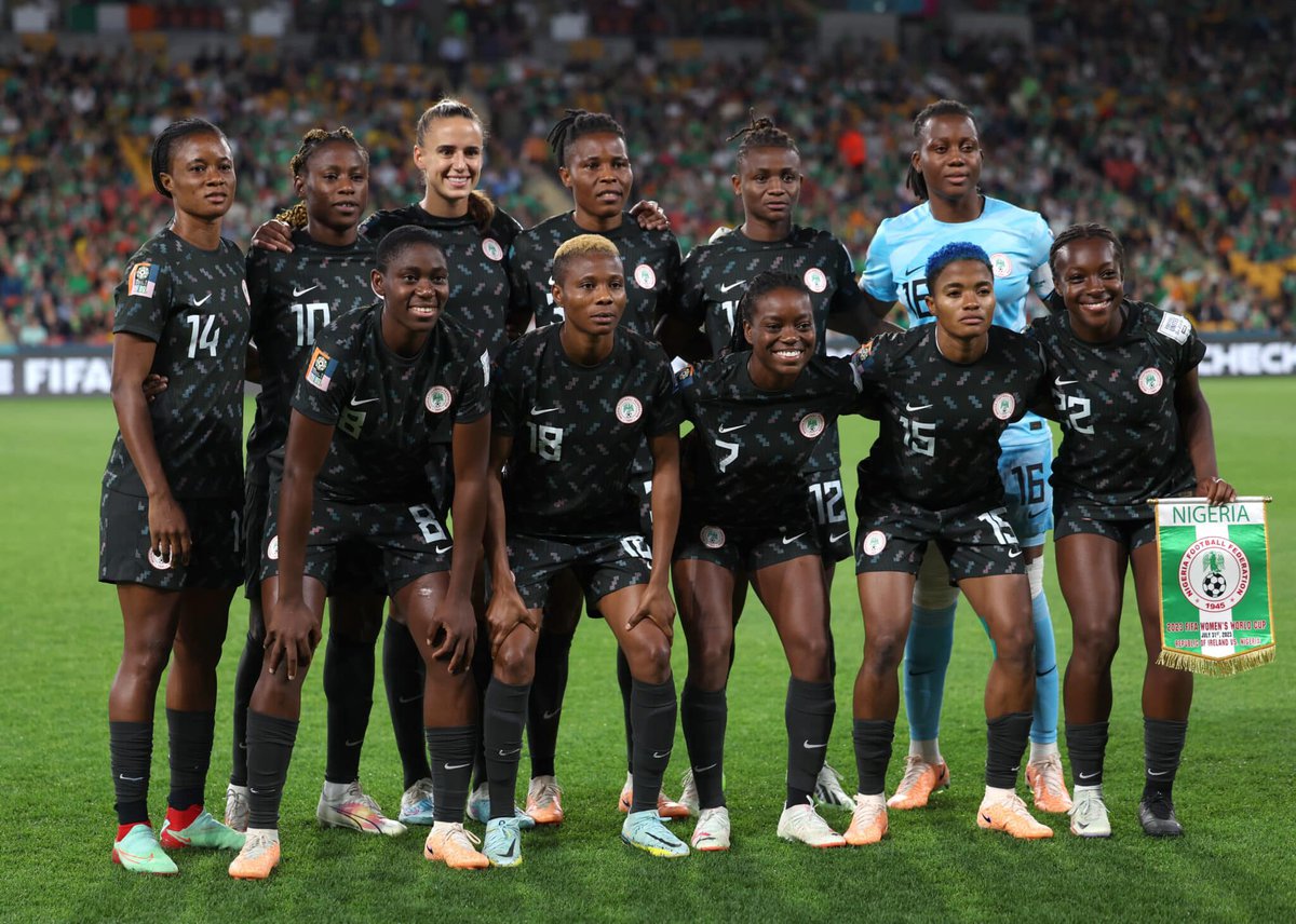 Nigeria women's national football team posing for a team photo before a match, wearing black jerseys with Nigerian flag patches, on a football pitch with a large stadium audience in the background.