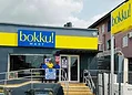 Brightly coloured storefront of Bokku Mart, a new convenience store serving the local community, with staff members standing at the entrance, showcasing a friendly shopping environment.