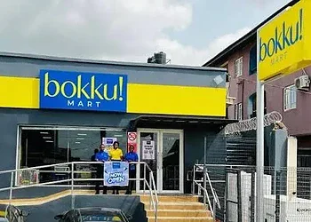 Brightly coloured storefront of Bokku Mart, a new convenience store serving the local community, with staff members standing at the entrance, showcasing a friendly shopping environment.