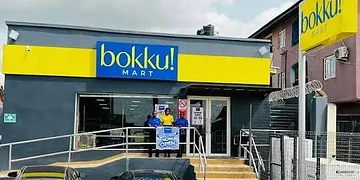 Brightly coloured storefront of Bokku Mart, a new convenience store serving the local community, with staff members standing at the entrance, showcasing a friendly shopping environment.