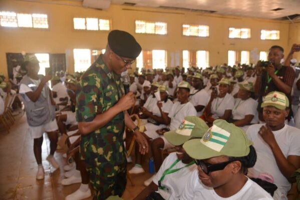 A young military officer speaks passionately to a diverse group of youth attendees at an indoor event, emphasizing skills and empowerment. The room is filled with attentive young people wearing caps and casual clothing, fostering a sense of community and leadership.