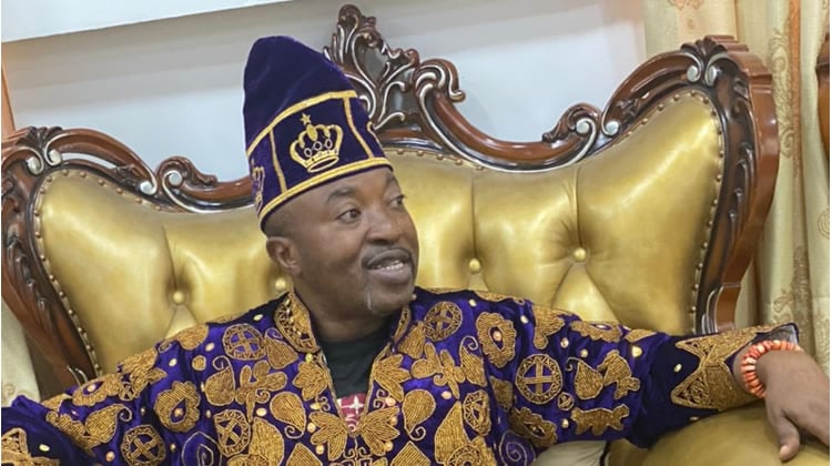 1. Man dressed in traditional African attire with intricate gold patterns and a matching cap, sitting on an ornate gold and wood sofa.