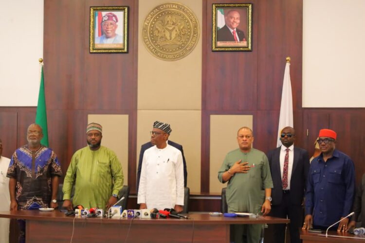 Senior Nigerian government officials during a formal meeting in the federal capital territory of Nigeria. The display includes photos of national leaders, national flags, and a gold emblem of Nigeria on the wall.