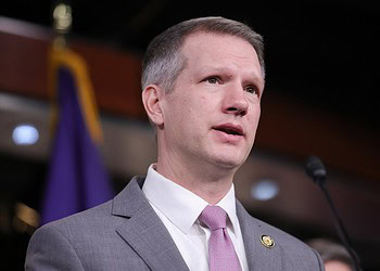 Alternative text: Close-up of a man in a grey suit with pink tie speaking at a press conference, with a microphone and purple background.