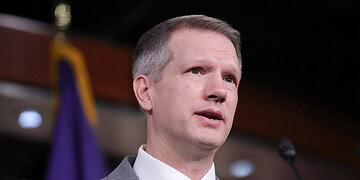 Alternative text: Close-up of a man in a grey suit with pink tie speaking at a press conference, with a microphone and purple background.