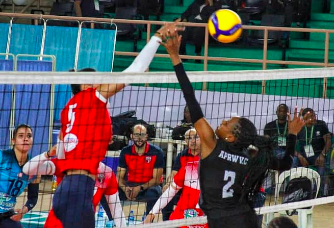 High-action volleyball match showing players at the net during a professional game, with spectators and officials in the background.