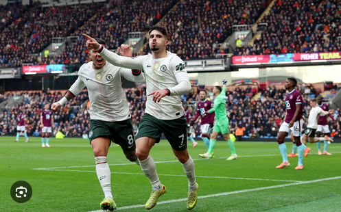 Two football players celebrating a goal on the field during a match at a packed stadium, football fans in the background, bright lights, and team uniforms in action.
