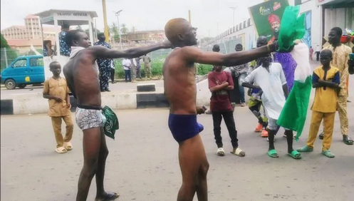 A group of barefoot boys performing a traditional dance on the street, with onlookers, cars, and a cityscape in the background, capturing cultural expression and community spirit.