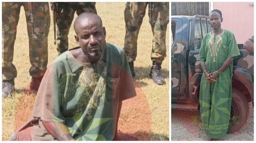 A man in traditional green attire standing beside a military vehicle, with soldiers in uniform in the background, during daytime outdoor setting.