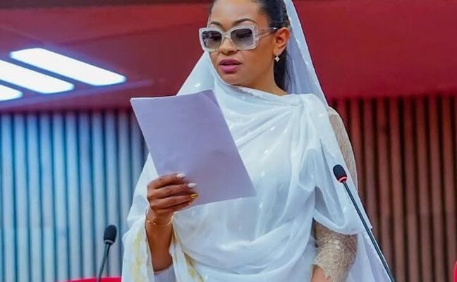 A woman in a white traditional dress and sunglasses reading a document at a formal event, with a red background and microphones, representing professionalism and cultural attire.